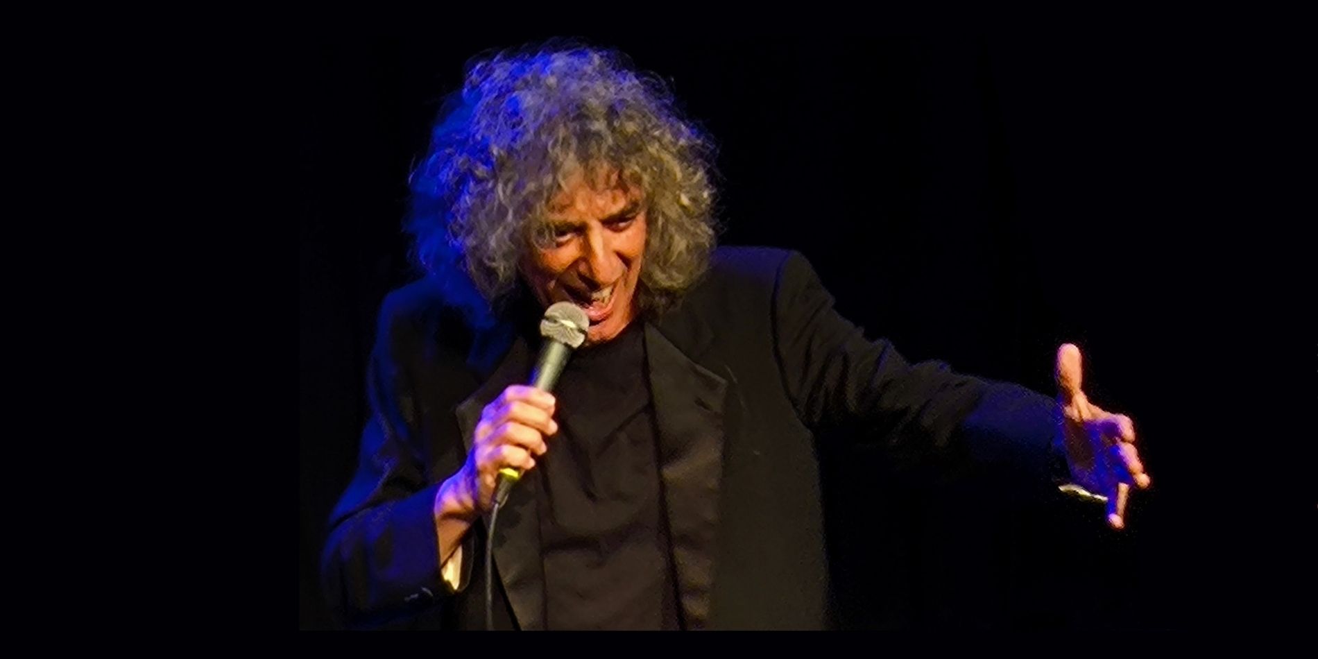 אישה עומדת על במה חשוכה, אוחזת במיקרופון ושרה בעוצמה, כשידה השנייה נשלחת קדימה בתנועה דרמטית. התאורה הממוקדת מדגישה את הבעתה ואת הנוכחות הבימתית הסוחפת.