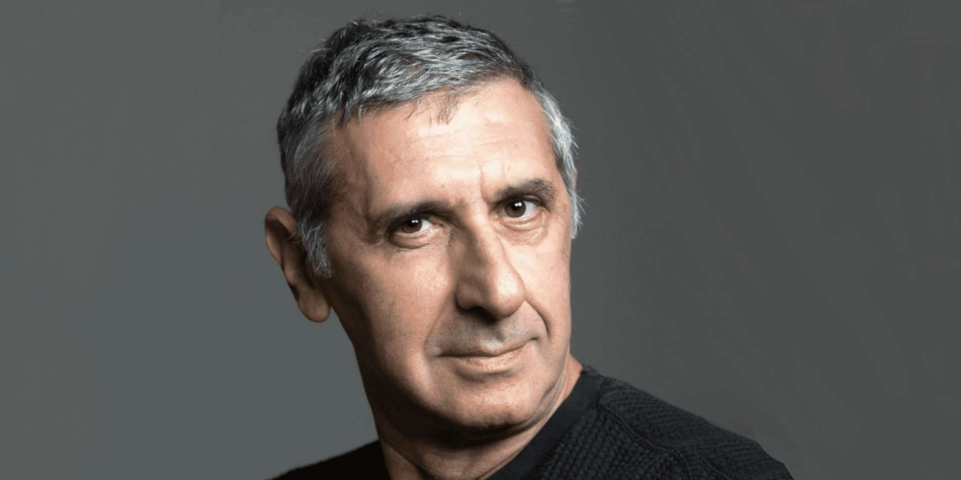 דיוקן תקריב של גבר מבוגר עם שיער מאפיר והבעה מהורהרת, מבטו מופנה מעט הצידה על רקע אחיד ואפור. התאורה הרכה מדגישה את תווי הפנים ויוצרת תחושה של איפוק ועומק.