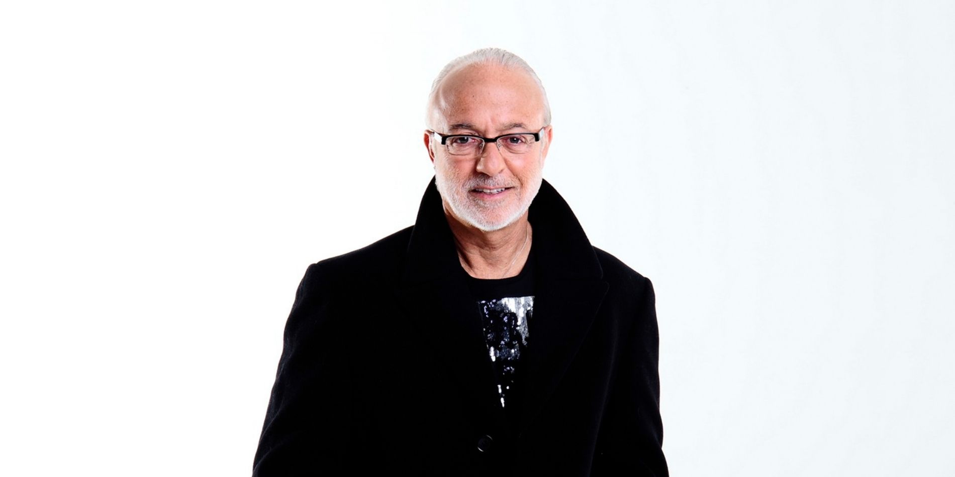 דיוקן של גבר מבוגר על רקע לבן, לבוש מעיל כהה ומשקפיים, מבטו ישיר ונינוח אל המצלמה. ההבעה רגועה ומאופקת, והצילום נקי וממוקד בפנים ובנוכחות האישית.