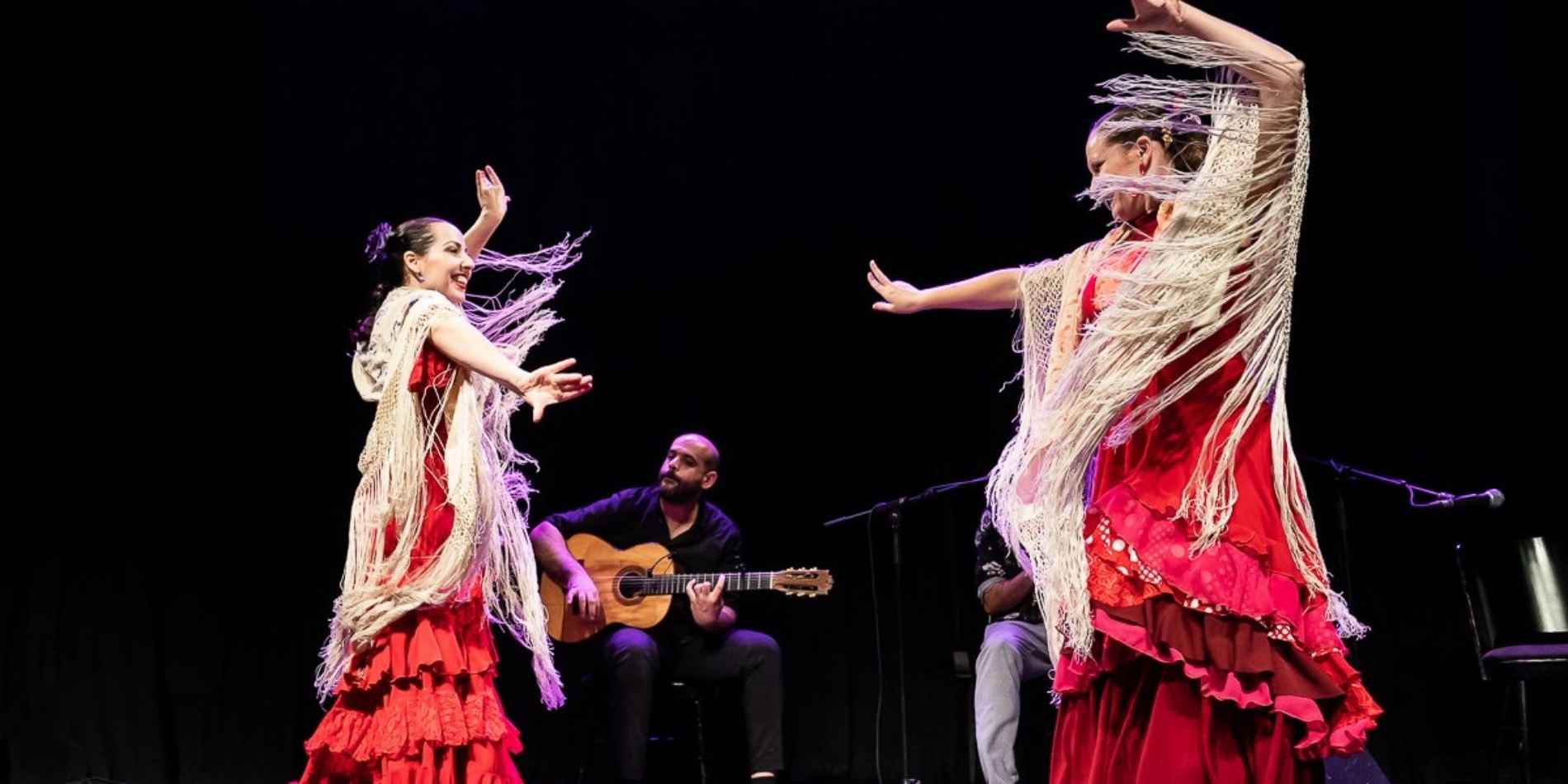 שתי רקדניות פלמנקו בשמלות אדומות ניצבות זו מול זו על הבמה, תנועות ידיהן חדות ומלאות הבעה, ושולי הצעיפים מתנפנפים סביבן. מאחוריהן יושב גיטריסט ומלווה את הריקוד בנגינה חיה, באווירה אינטימית ועוצמתית.
