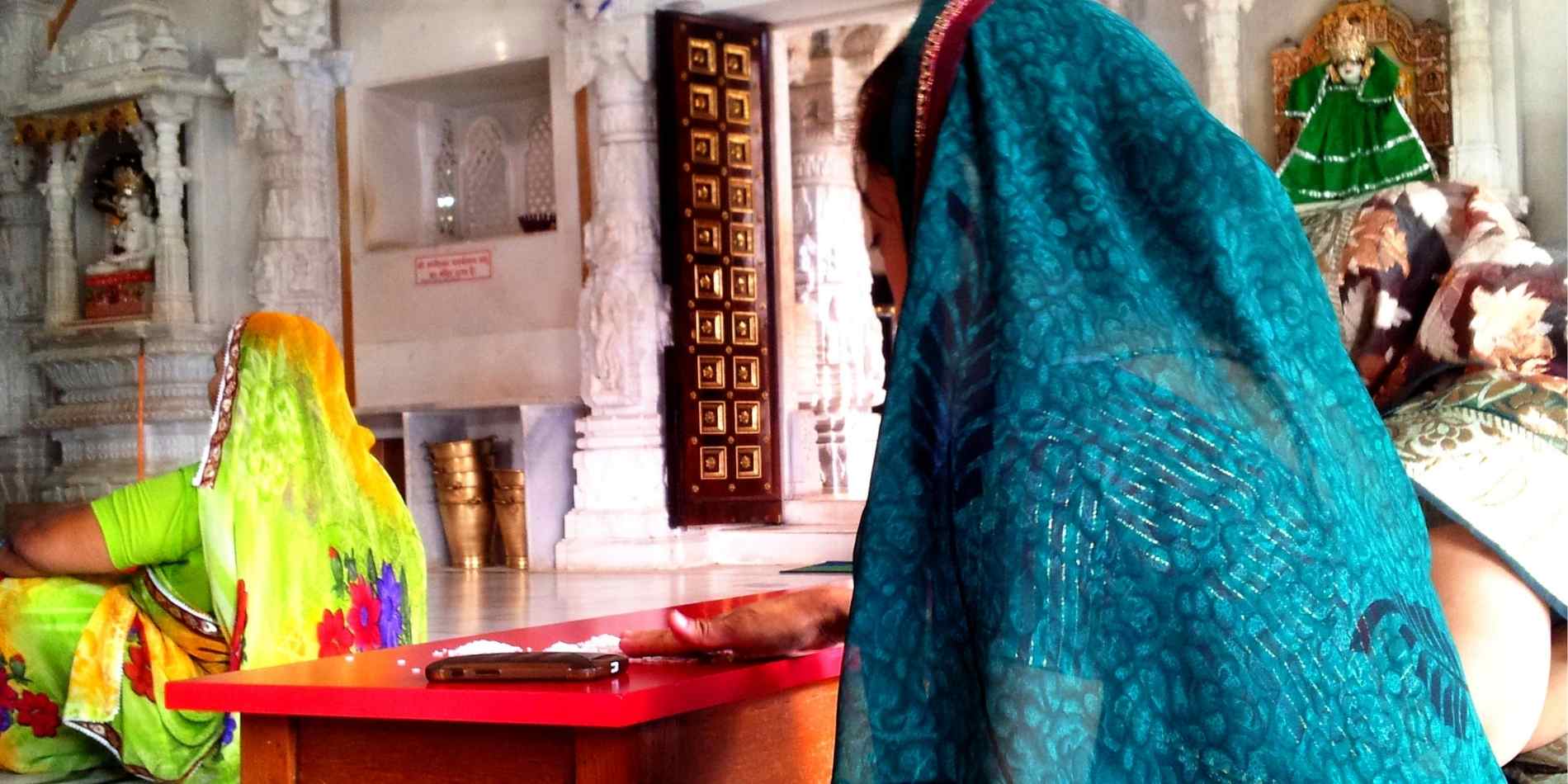 אישה עטויה סארי כחול נשענת על שולחן אדום בתוך מקדש שיש, ברגע שקט של התכנסות והתבוננות, כשסביבה דמויות נוספות בלבוש מסורתי. המפגש בין היומיום, הגוף והמרחב הקדוש יוצר תחושת אינטימיות ושגרה רוחנית.