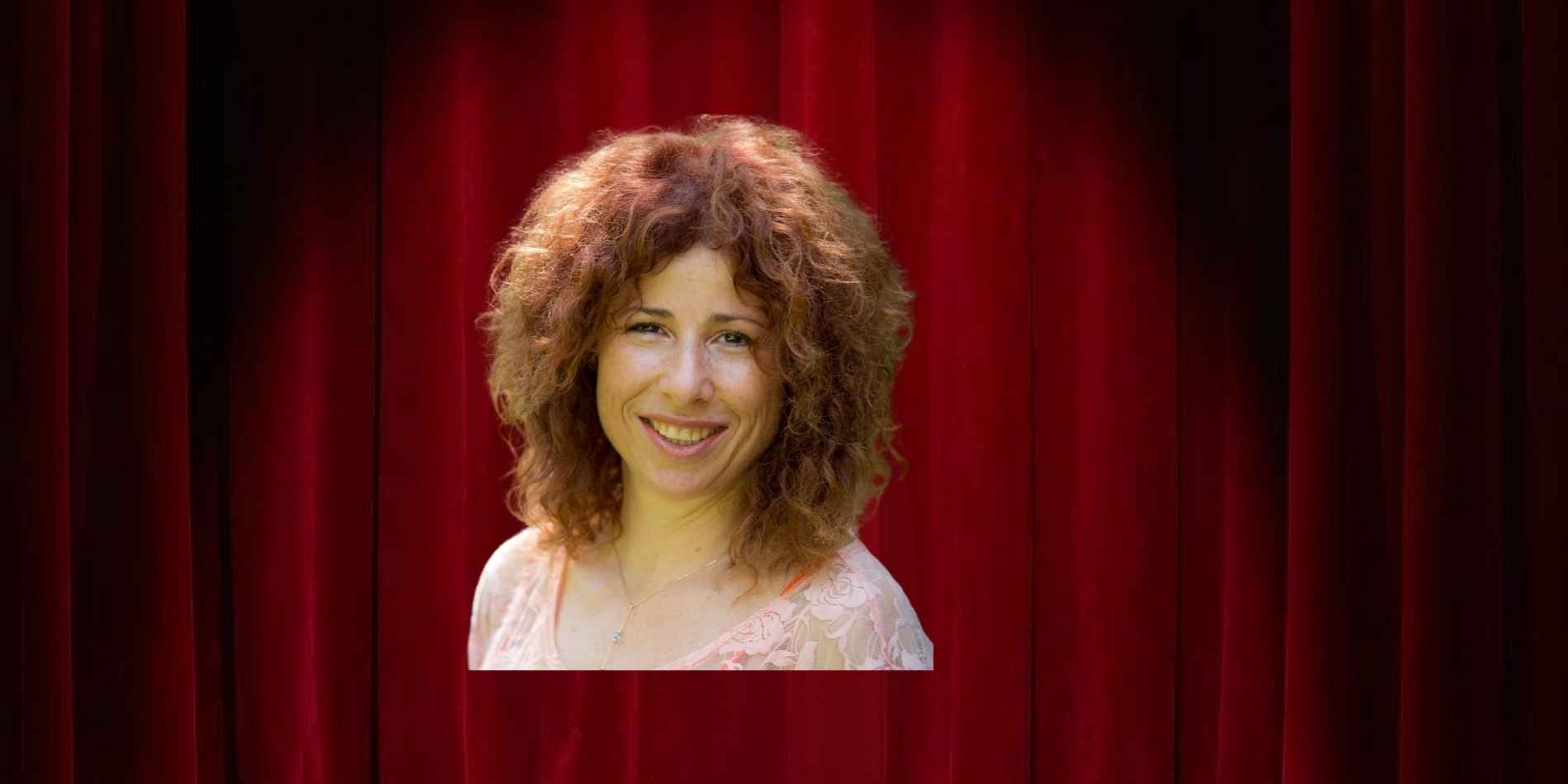 דיוקן צבעוני של אישה צעירה עם שיער מתולתל בגוון אדמוני. היא מחייכת ישירות אל המצלמה, לבושה חולצה בהירה ועדינה, על רקע כהה ואחיד שמבליט את פניה והבעתה. התאורה רכה וטבעית ויוצרת תחושה פתוחה, נעימה ומזמינה.