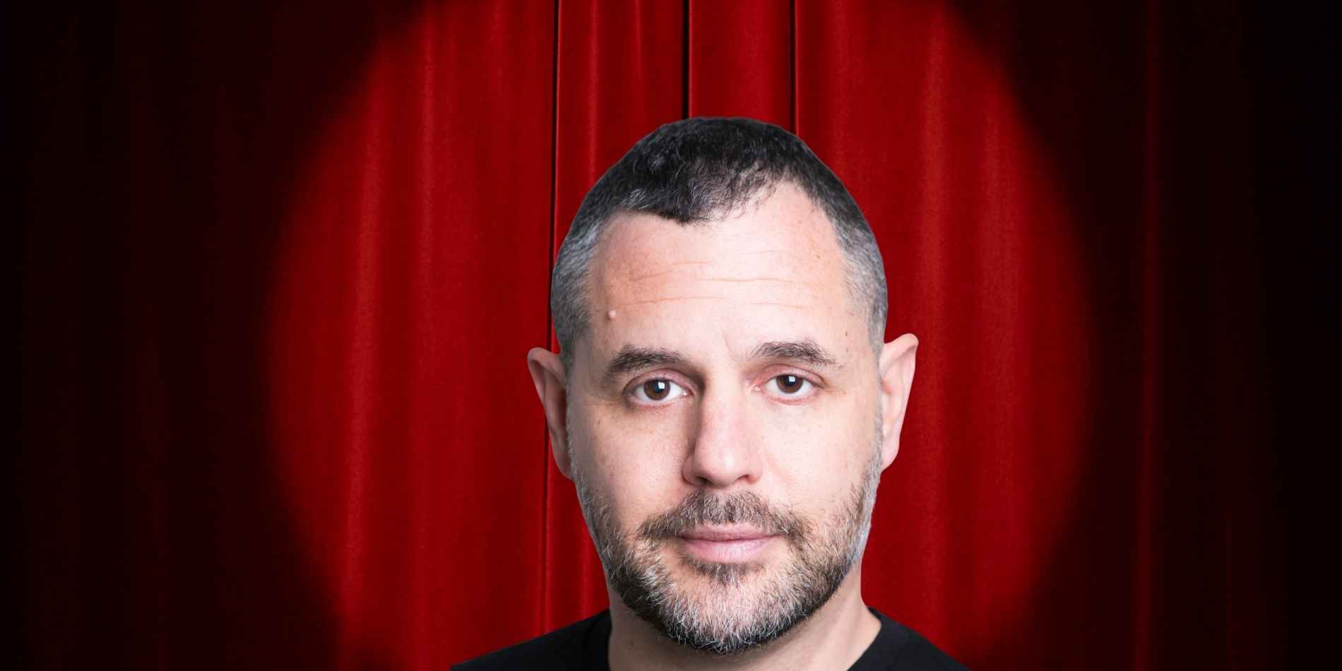 בתמונה נראה גבר בגיל בוגר, מצולם בצילום סטודיו חזיתי. פניו במרכז הפריים, מבטו ישיר אל המצלמה והבעתו רגועה ונייטרלית. שיערו קצר מאוד, כהה עם התחלה של שיבה, ויש לו זקן קצר ומטופח. הוא לובש חולצה כהה וחלקה ללא דוגמה. הרקע הוא וילון אדום עמוק, המזכיר במה תיאטרונית או מסך קטיפה, ויוצר קונטרסט ברור עם גוון העור והלבוש. התאורה רכה ומאוזנת, מדגישה את תווי הפנים ללא צללים חדים, ונותנת לתמונה אופי רשמי ומוקפד.