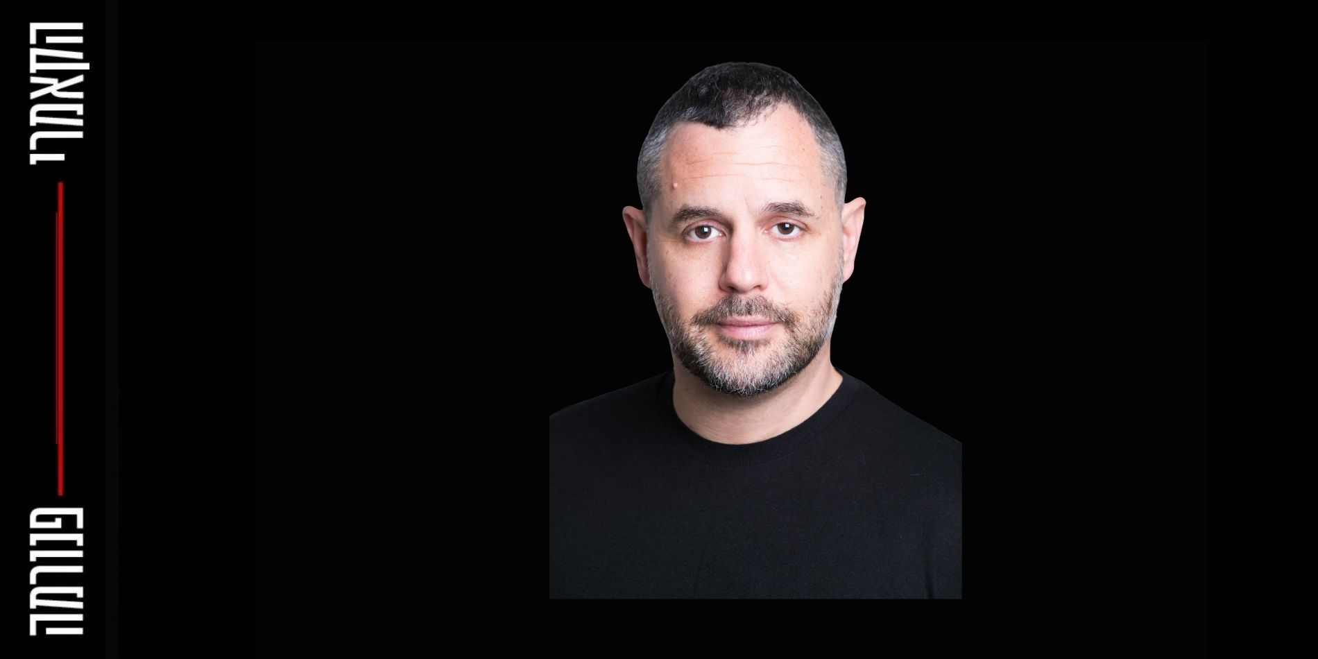 בתמונה נראה גבר מצולם בצילום סטודיו חזיתי. פניו במרכז הפריים, מבטו ישיר אל המצלמה והבעתו רגועה ונייטרלית. שיערו קצר מאוד, כהה עם התחלה של שיבה, ויש לו זקן קצר ומטופח. הוא לובש חולצה כהה וחלקה ללא דוגמה. הרקע הוא וילון אדום עמוק, המזכיר במה תיאטרונית או מסך קטיפה, ויוצר קונטרסט ברור עם גוון העור והלבוש. התאורה רכה ומאוזנת, מדגישה את תווי הפנים ללא צללים חדים, ונותנת לתמונה אופי רשמי ומוקפד.