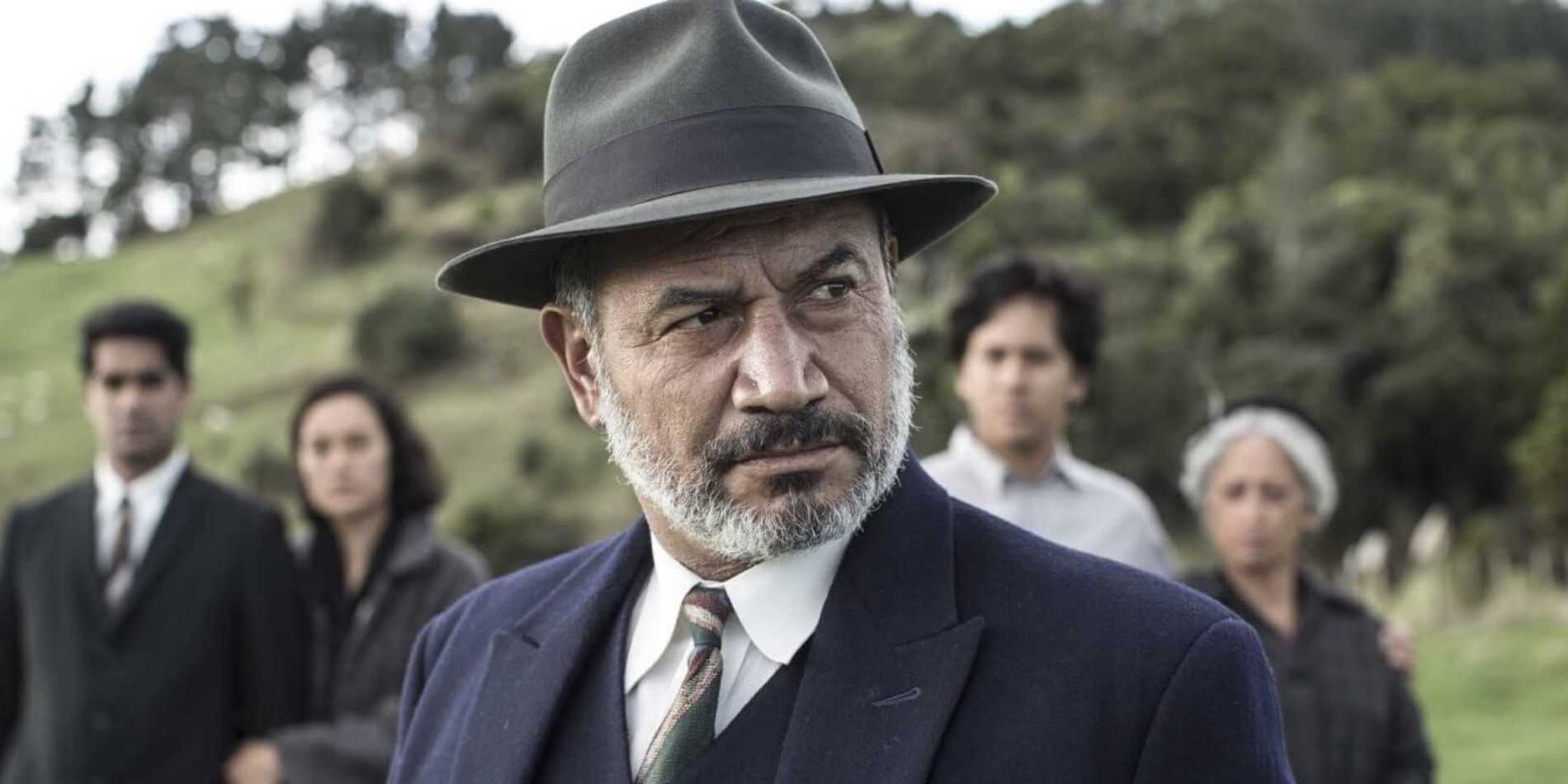 בתמונה נראה גבר מבוגר עומד בקדמת הפריים, לבוש חליפה כהה בסגנון מוקפד וכובע רחב שוליים. יש לו זקן ושיער מאפיר, והבעתו רצינית, דרוכה ומהורהרת. הוא מפנה את מבטו הצִדה, כאילו מגיב למשהו שמתרחש מחוץ לפריים, ומשדר סמכות ונוכחות חזקה. מאחוריו, במרחק קצר ובטשטוש קל של עומק שדה, עומדת קבוצה של ארבעה אנשים – גברים ונשים – לבושים בבגדים פשוטים וכהים יחסית. הם ניצבים יחד, בשקט, ומבטיהם מופנים לכיוונו או לאותו כיוון שאליו הוא מסתכל, מה שיוצר תחושת מתח וציפייה. הרקע הוא נוף פתוח וטבעי: גבעות ירוקות, עצים וצמחייה, באור יום רך. האווירה הכללית דרמטית ומאופקת, עם תחושה של סיפור היסטורי או משפחתי, מנהיגות, אחריות וקונפליקט סמוי.