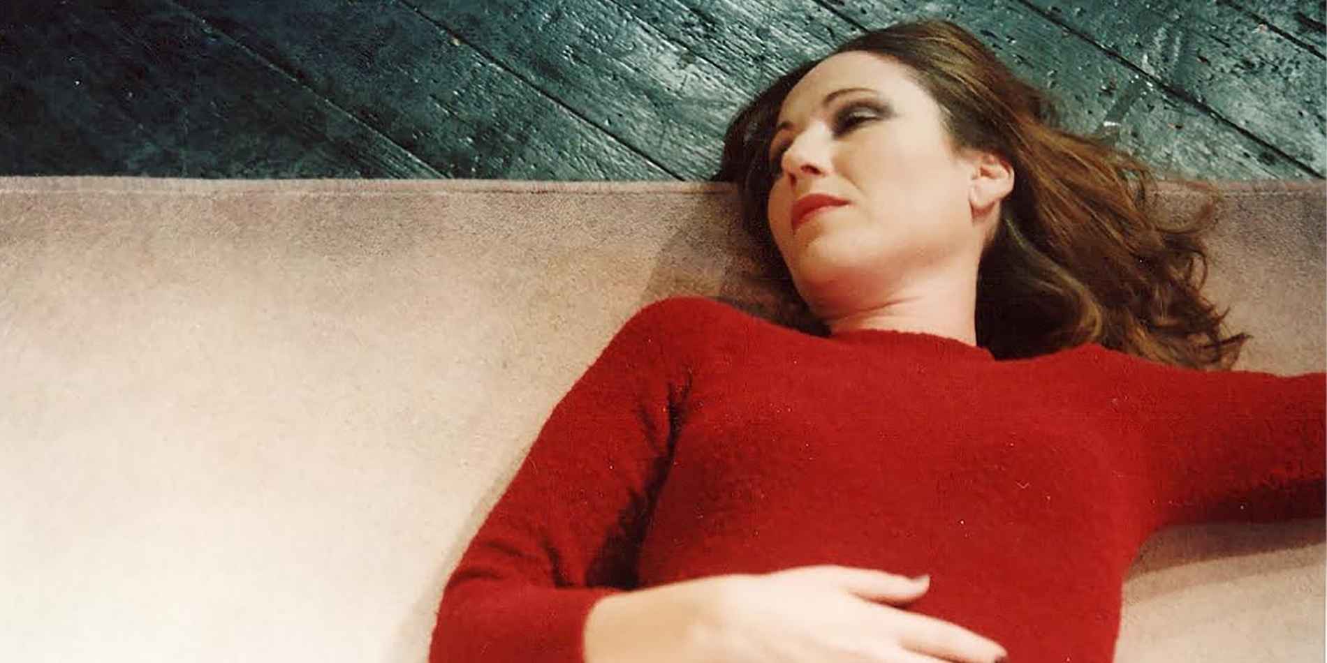 אישה שוכבת על ספה בהירה, לבושה סוודר אדום, מבטה מופנה הצידה כאילו שקועה במחשבה או בזיכרון רחוק. הניגוד בין האדום החם לגוונים הקרים של הרצפה והסביבה יוצר תחושת שקט טעון, בין מנוחה, פגיעוּת והמתנה.