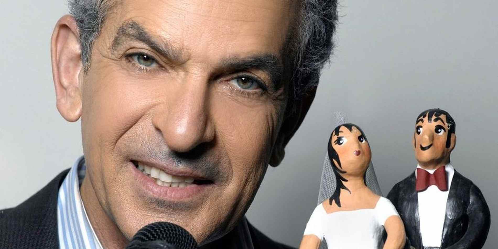 פורטרט רחב שלגבר עם מיקרופון בצד שמאל של הפריים, כשמימין מאחוריו ניצבים בבירור פסלוני חתן־כלה בסגנון מצויר. הרקע האפור והמרווח סביב הדמויות נותנים תחושת סטודיו רגועה עם מוקד כפול: האדם בקדמה והפסלונים ברקע