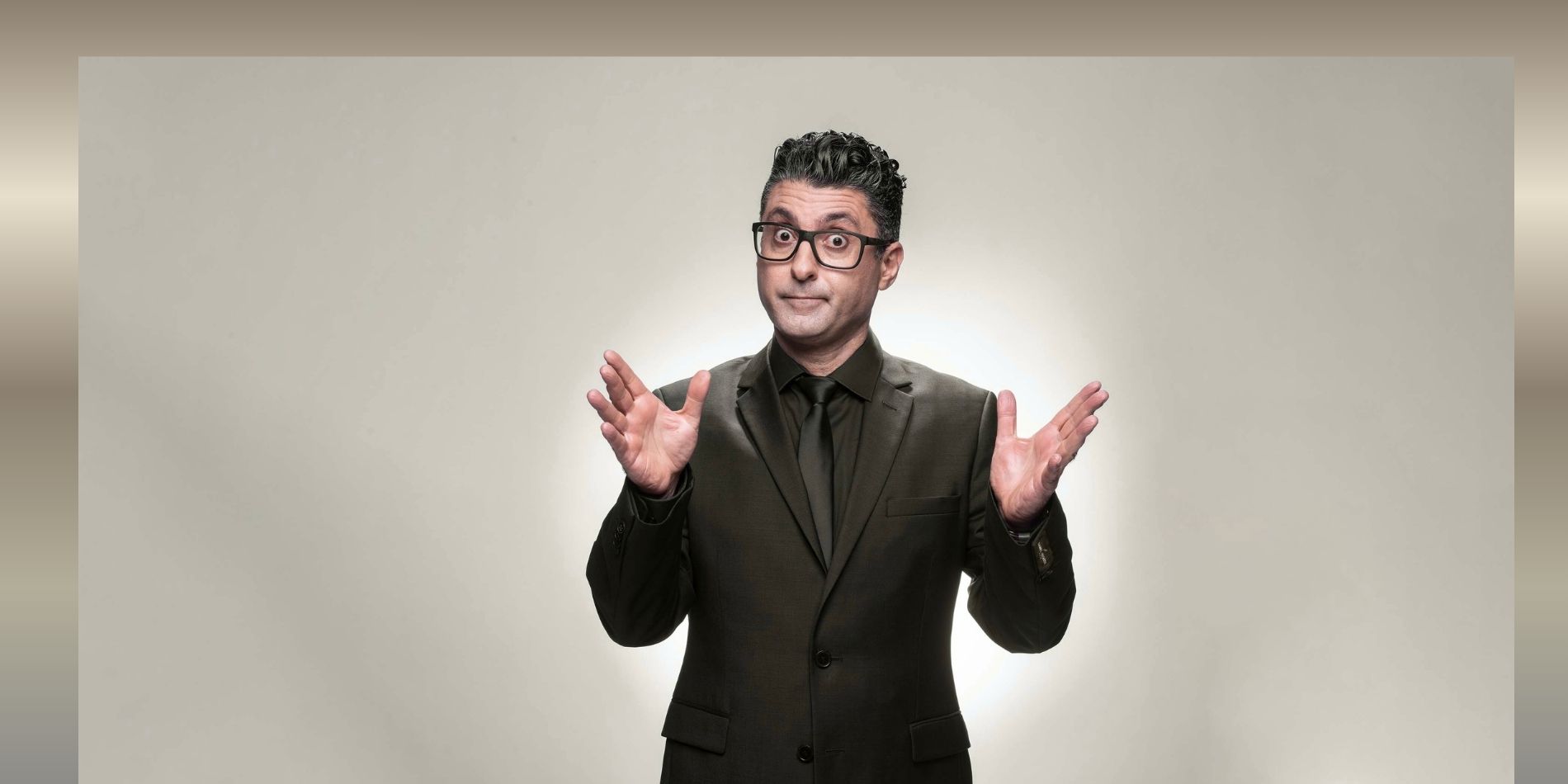 דמות גברית בחליפה כהה ומשקפיים עומדת מול רקע בהיר ונקי, מביטה ישירות למצלמה בהבעה חדה ומלאת הומור מאופק. הידיים מורמות בתנועה פתוחה ומדויקת, היוצרת תחושה של פנייה אל הקהל, שילוב בין סמכות, אירוניה ונוכחות בימתית.