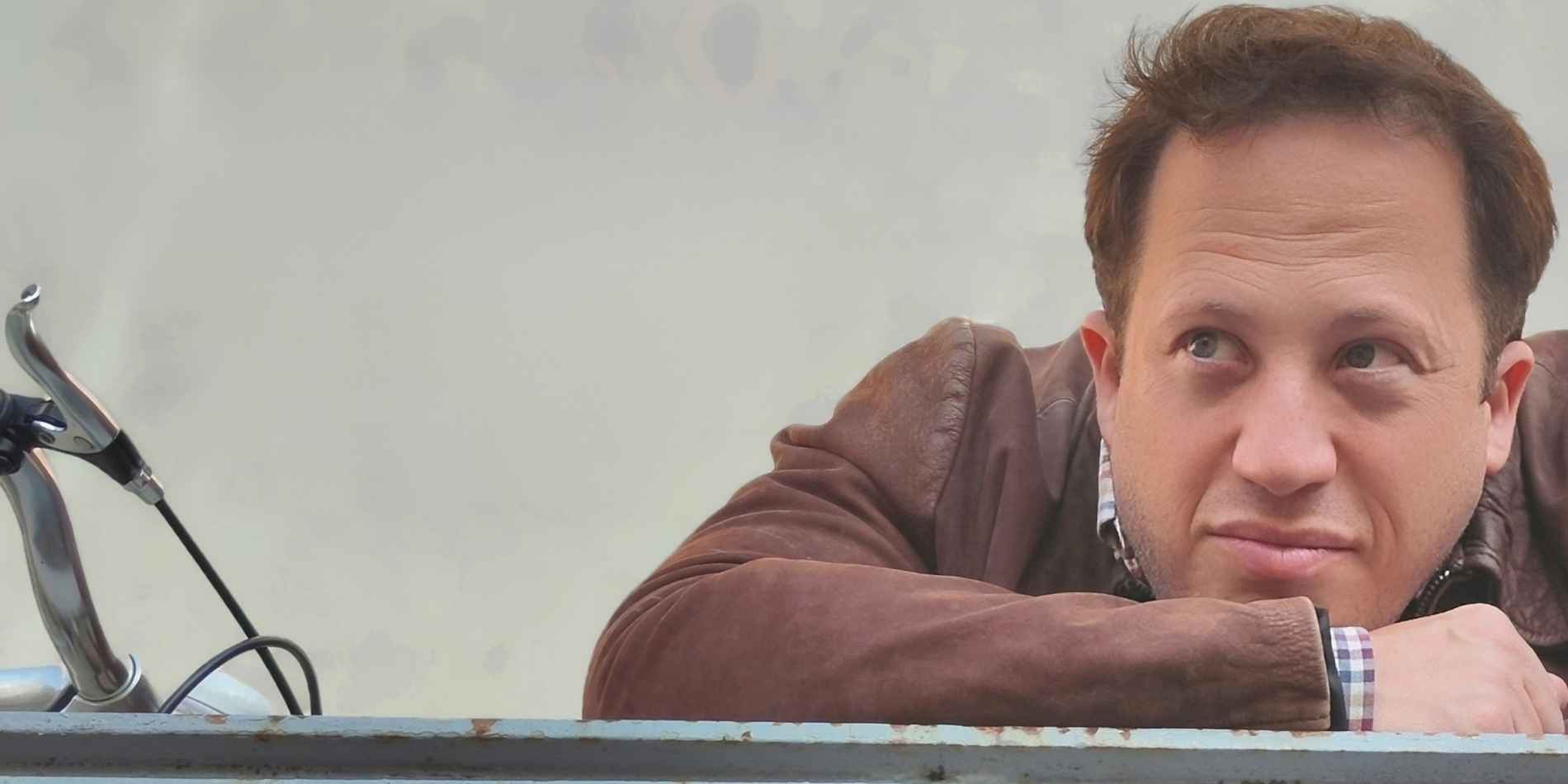 גבר נשען על מעקה מתכת, זרועותיו משולבות ומבטו מופנה הצִדה בהבעה מהורהרת וקצת ספקנית. הרקע הבהיר והאופניים שבשוליים יוצרים רגע יומיומי שקט, בין תנועה לעצירה.