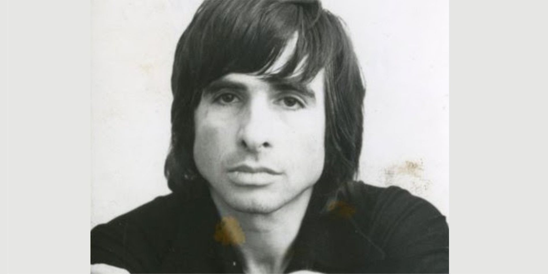 דיוקן שחור־לבן של גבר צעיר במבט ישיר אל המצלמה, שיער כהה וארוך נופל ברכות על מצחו ופניו רזות ומהורהרות. התאורה הרכה והרקע הבהיר מדגישים הבעה שקטה ואינטימית, כמעט פגיעה.