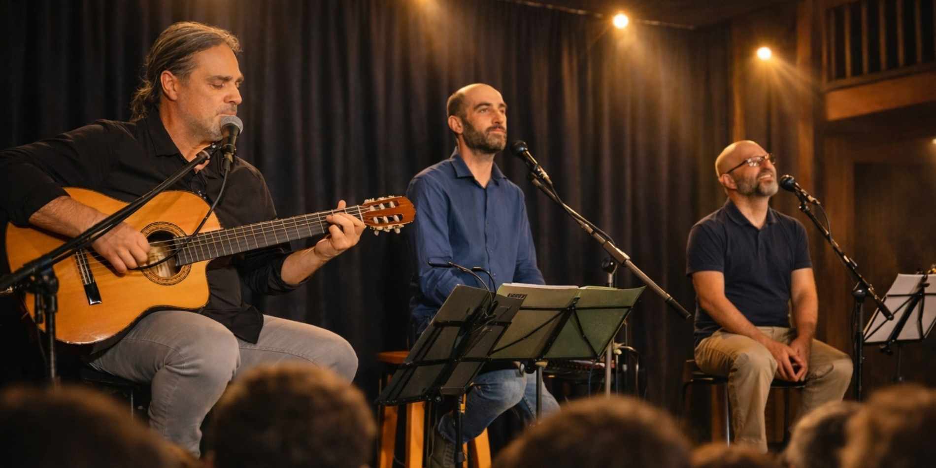 שלושה מוזיקאים יושבים על במה קטנה מול קהל: אחד מנגן בגיטרה אקוסטית ושר, ושני האחרים שרים למיקרופונים לצד עמודי תווים. התאורה החמה והווילון הכהה ברקע יוצרים אווירה אינטימית של מופע מוזיקלי חי.